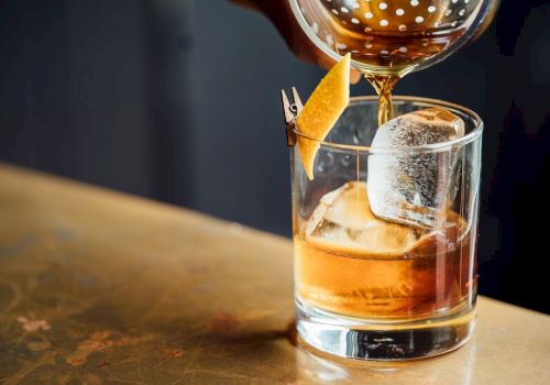 A whiskey glass with ice and a garnish being poured, a stylish drink on a wooden bar. (Note: The sentence ends with a period.)