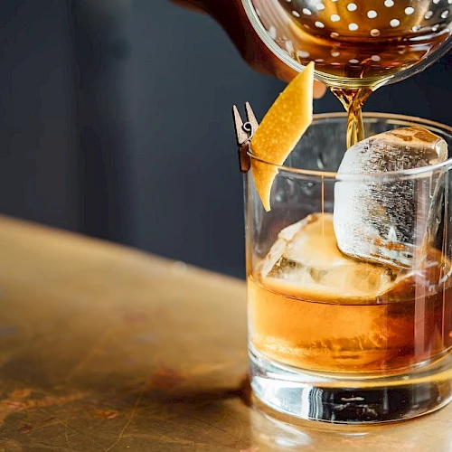 A whiskey glass with ice and a garnish being poured, a stylish drink on a wooden bar. (Note: The sentence ends with a period.)