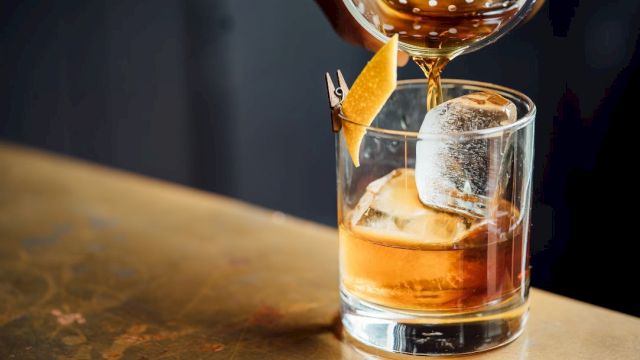 A whiskey glass with ice and a garnish being poured, a stylish drink on a wooden bar. (Note: The sentence ends with a period.)