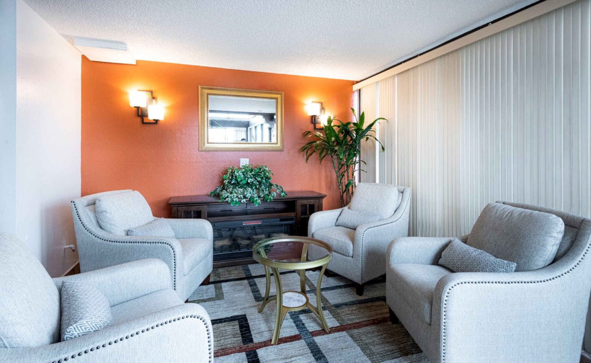 A cozy waiting room with four light gray armchairs around a small glass table, orange accent wall, and plants.