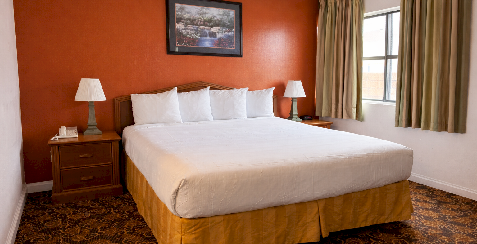 A tidy hotel room with a large bed, white linens, two bedside tables with lamps, a red accent wall, and a window with curtains.