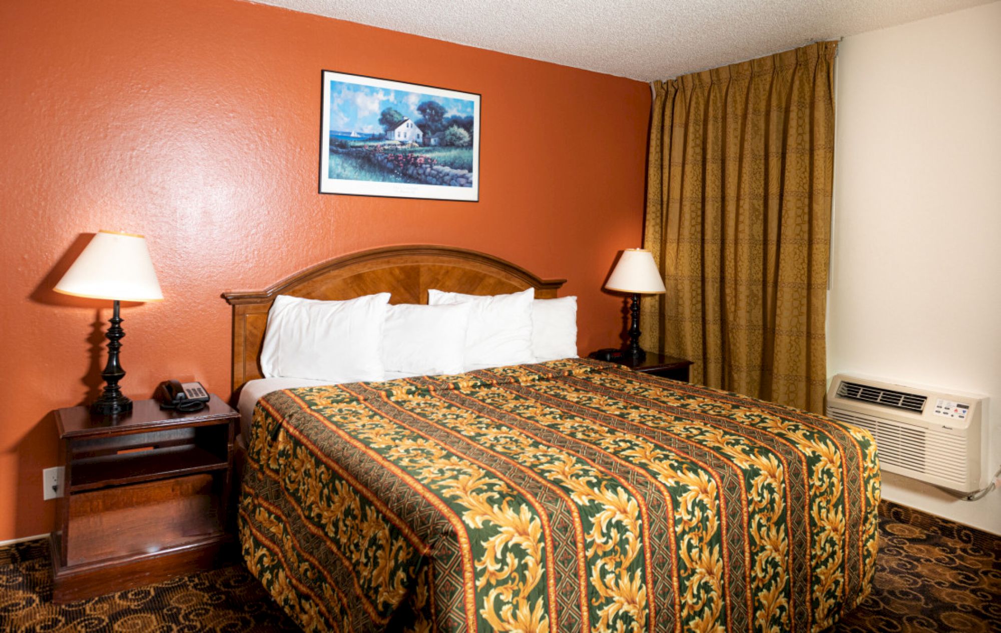 A hotel room with a king bed, orange accent wall, two bedside lamps, framed artwork, heavy curtains, and a window air conditioner.