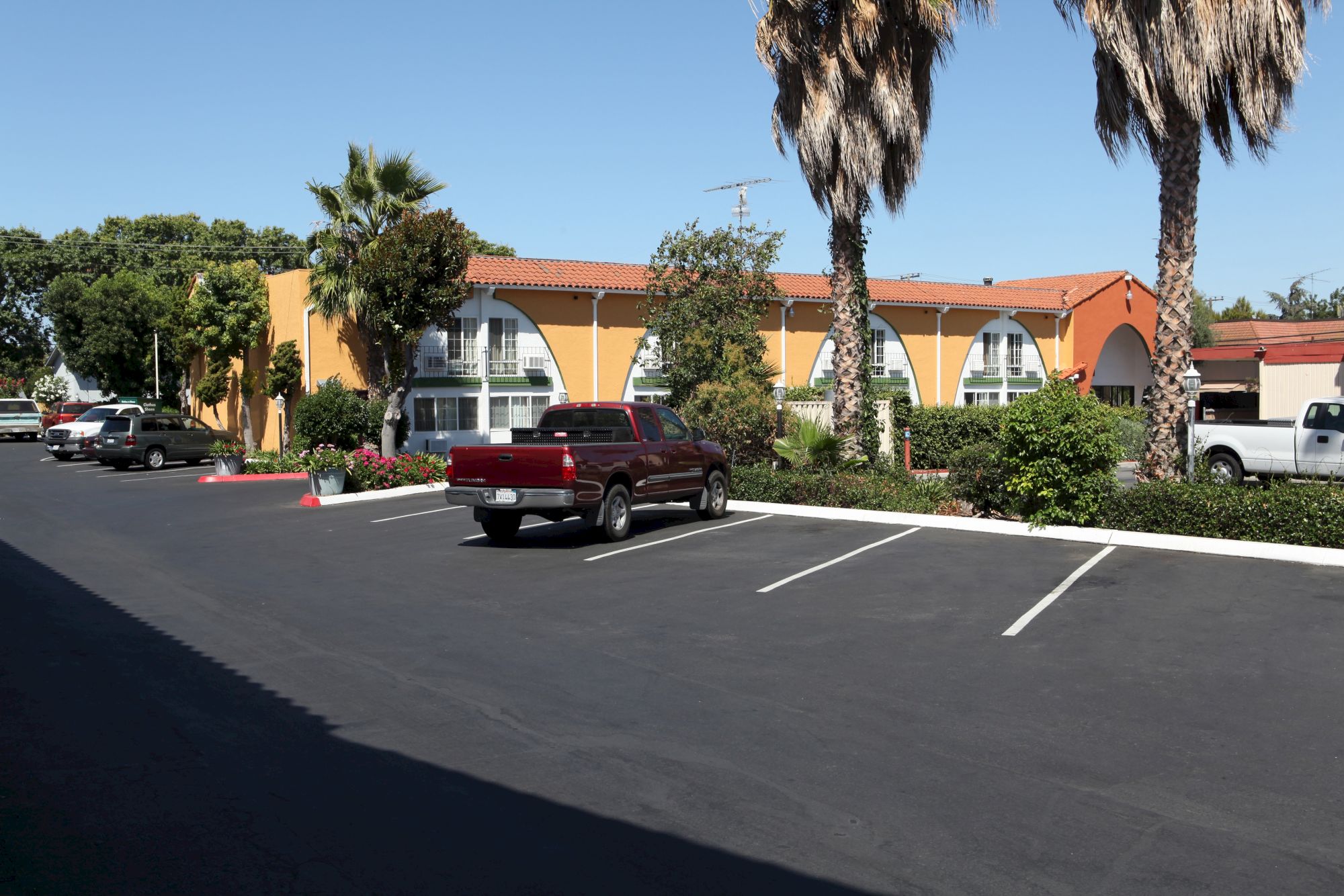 A parking lot in front of a sunny orange building with palm trees; a red pickup is parked, and white lines mark spaces for cars.