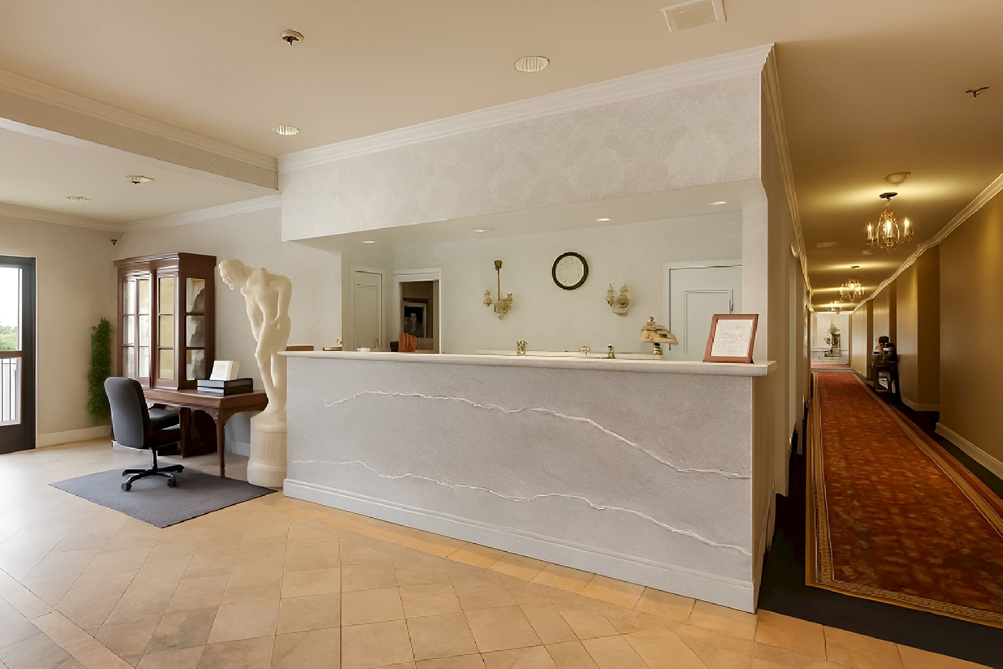 A hotel lobby with a marble-front desk, a desk chair by a small piano, hallway carpet, and warm lighting.
