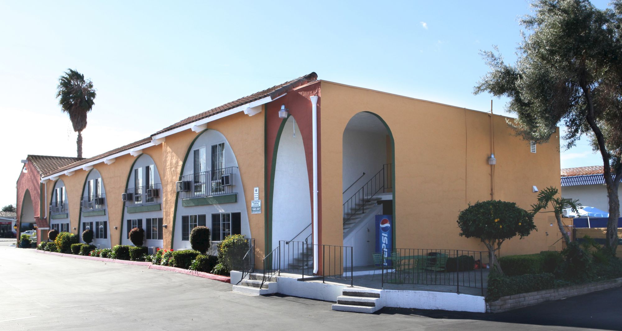 A colorful, arched-row motel or apartment building with yellow walls, arched doorways, blue trim, and a few palm trees at the corner, sunny day.