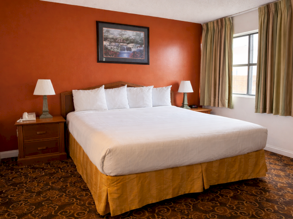 A tidy hotel room with a king bed, two nightstands, lamps, a framed picture, and a red accent wall by a window.