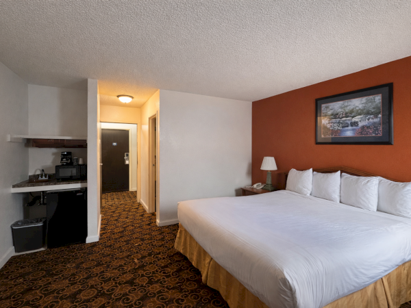 Hotel room with a large bed against a red accent wall, white linens, nightstand with lamp, a small desk/kitchenette area, and patterned carpet. End.