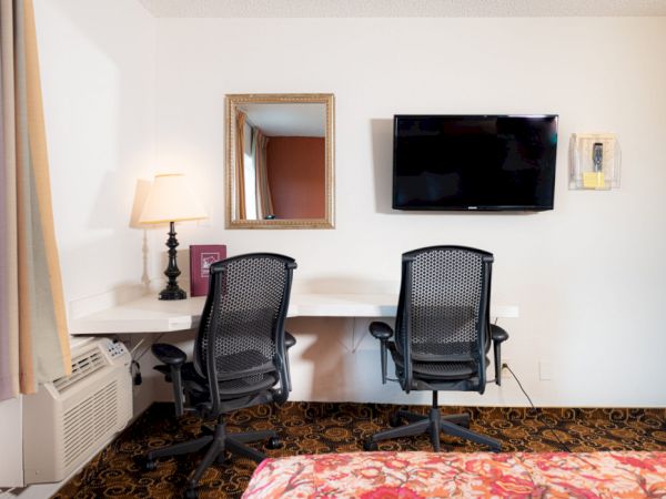 A hotel desk with two chairs, a wall-mounted TV, a mirror, a lamp on the desk, an air conditioner unit, and a portion of a floral bedspread in the foreground.
