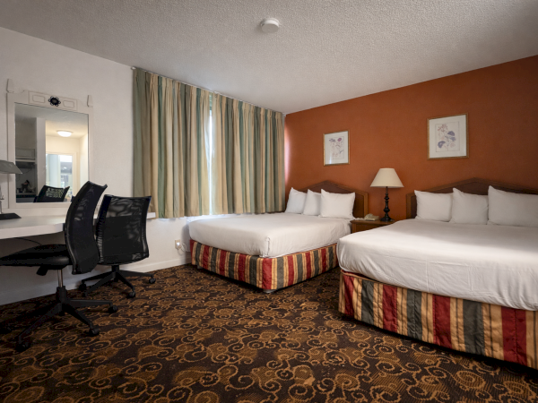 A hotel room with two queen beds, striped bed skirts, a desk and chair, a mirror, a lamp, and a window with turquoise curtains. A patterned carpet covers the floor.