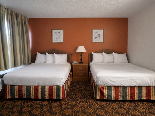 Two double beds with white linens, a nightstand, lamp, framed art, and a patterned carpet in a hotel room with a red accent wall.