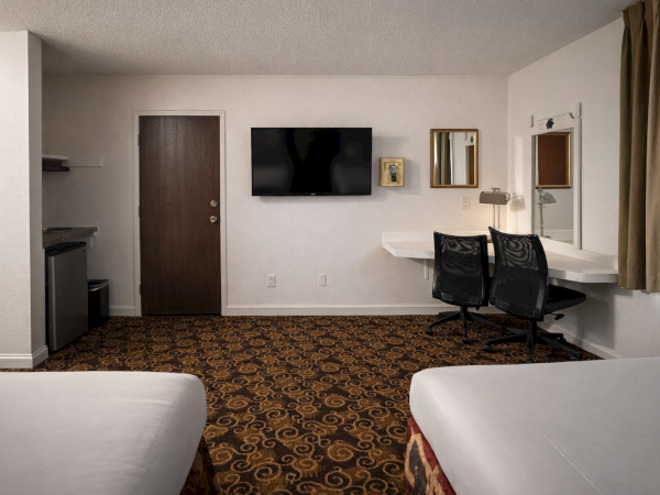 A hotel room with two beds, a wall TV, a desk with two chairs, a sink area, and a door, all in a simple, tidy layout.