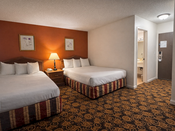 Two double beds with white linens, a red accent wall, nightstand and lamp, patterned carpet, and a doorway to a bathroom in a hotel room.