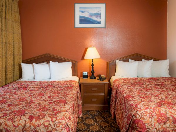 Two double beds with red floral bedspreads in a hotel room, a nightstand with a lamp between them, and a framed picture above the headboard.