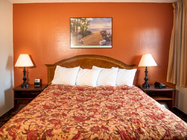 A warmly lit hotel room with a large bed, red-orange accent wall, floral bedspread, two nightstands with lamps, and a framed picture above.