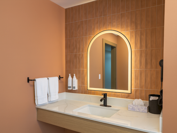 A stylish bathroom vanity with a backlit arched mirror, peach walls, tan tile backsplash, white countertop, towel rack, and soap dispensers.
