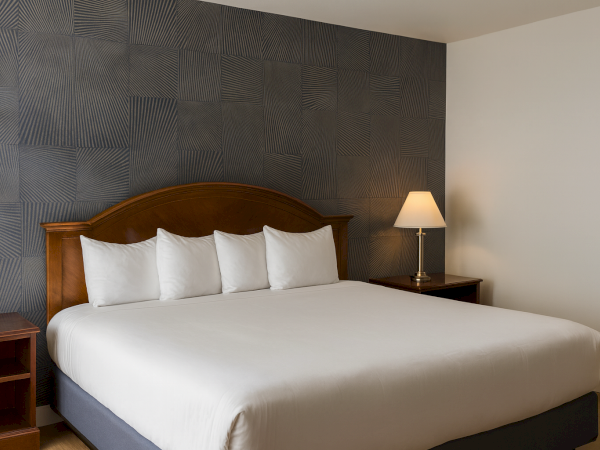 A tidy hotel room with a large bed, white linens, four pillows, dark wood headboard and nightstands, and a lamp on the right.