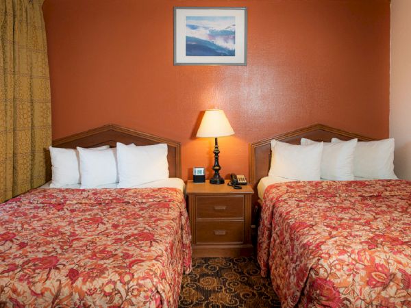 Two double beds with red floral bedspreads, a nightstand between them, a lamp, and a framed picture on a reddish wall.