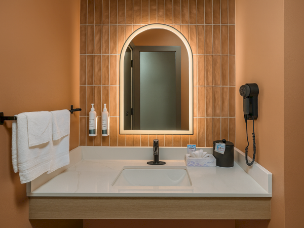 It&rsquo;s a modern bathroom sink vanity with an arched backlit mirror, peach-tiled wall, towel rack, and wall phone, neat and cozy.