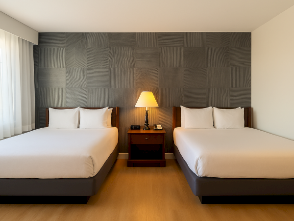 Two neatly made double beds with white linens, a shared nightstand, a lit table lamp, and a dark textured accent wall in a bright hotel room.