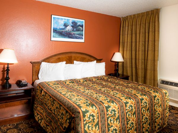 A hotel room with a king bed, red accent wall, two nightstands, lamps, framed art, curtains, and a wall AC unit. End.