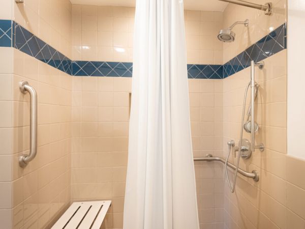 A tiled accessible shower with a curved curtain rod, white curtain, grab bars, a foldable bench, and a handheld showerhead for easy bathing.