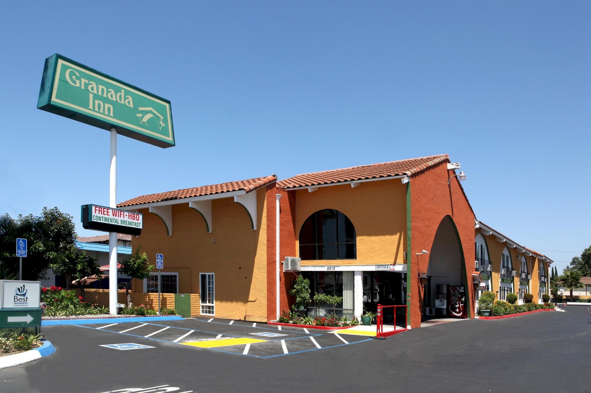 A Granada Inn with a tan and orange arched building, blue sky, and a green sign reading &ldquo;Granada Inn&rdquo; beside a parking lot.