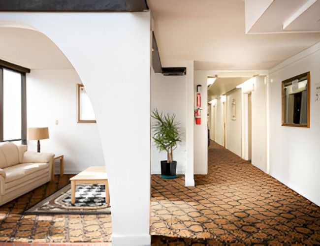 A hotel lobby with a seating area on the left, white walls, archways, a long carpeted hallway, potted plants, and a rack of brochures on the right.