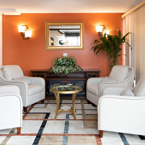 A cozy hotel lobby seating area with four white armchairs around a dark wood table, an orange accent wall, wall sconces, a plant, and a reflective mirror above a console.