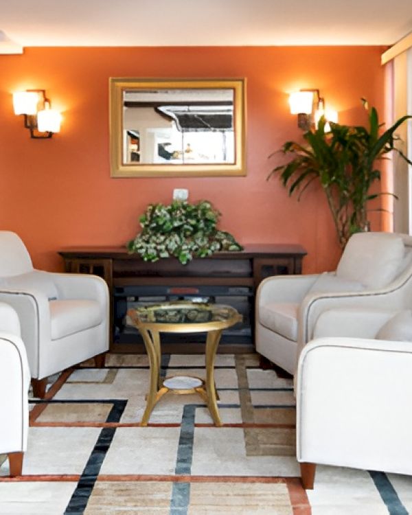 A cozy hotel lobby seating area with four white armchairs around a dark wood table, an orange accent wall, wall sconces, a plant, and a reflective mirror above a console.