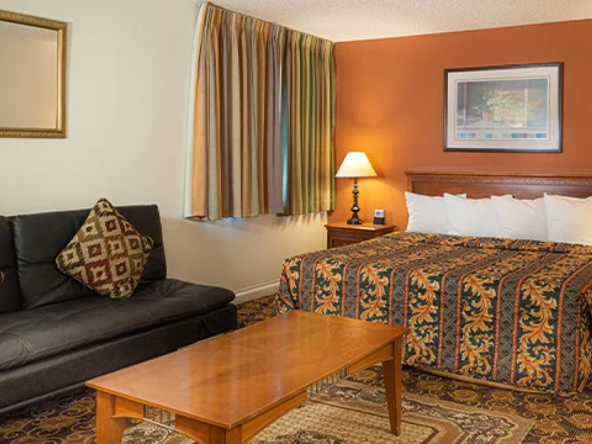 A hotel room with a king bed, two nightstands, matching lamps, a patterned bedspread, a sofa with decorative cushions, and a wooden coffee table.