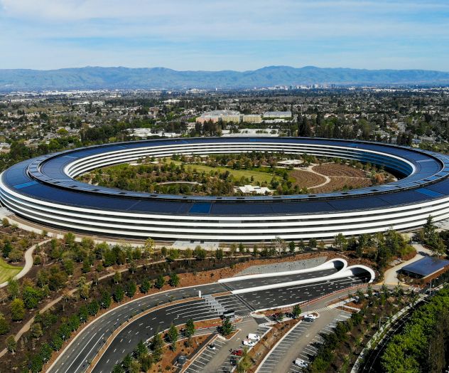Aerial view of a large circular building, the Apple Park-style ring, surrounded by roads, greenery, and a few parked cars, in a suburban landscape.