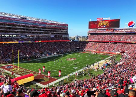 A packed football stadium with red-clad fans, a green field, players on the field, and a bright afternoon sky ending the scene.