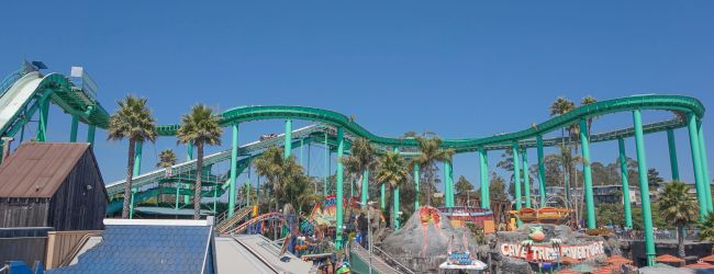 A sunny amusement park with a bright green roller coaster, palm trees, queues, and colorful shops and umbrellas along a crowded walkway.