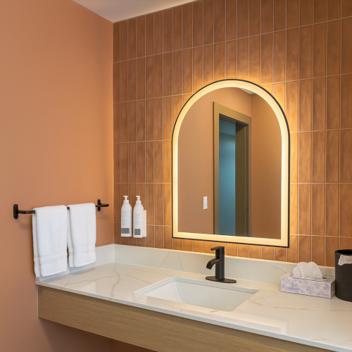 A modern bathroom with a floating vanity, beige walls, a backlit arched mirror, twin towel bars, and a white sink with minimalist accessories, ends.