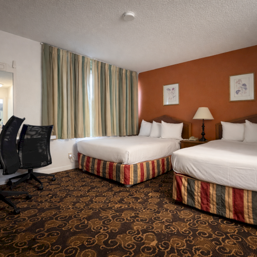 A hotel room with two single beds, a desk, chair, wall-mounted TV, and a bright orange accent wall, plus a window and ceiling light.