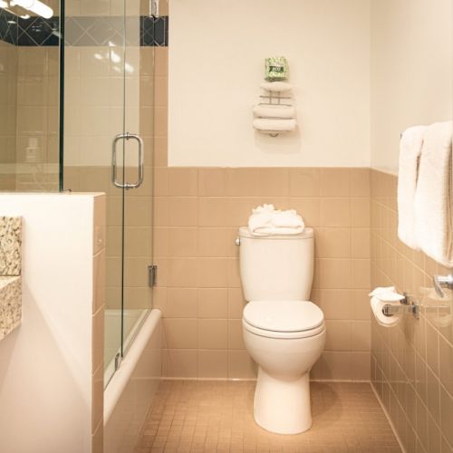 A clean, modern bathroom featuring a toilet with stacked towels, a glass shower enclosure, beige wall tiles, and a towel rack with a white towel.