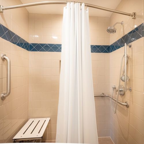 A beige-tiled bathroom with a white shower curtain, blue tile border, a wall-mounted shower with grab bars, and a small wooden seat.
