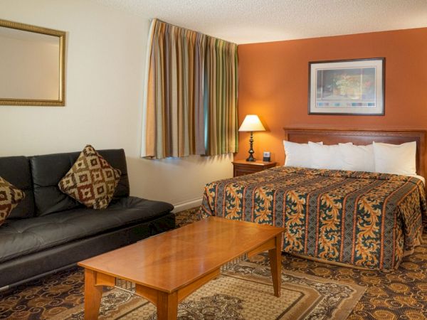 A hotel room with a king bed, two nightstands, lamps, a sofa, a wooden coffee table, patterned carpet, and warm orange accent wall. The bed has a quilted cover.