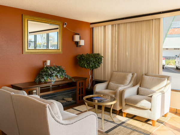 A cozy hotel lobby seating area with four beige cushioned chairs, a wooden console, potted plants, a wall mirror, and warm orange accent wall by a large window.